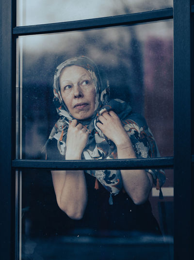Sad mature woman with scraft on head standing behind dirty window glass