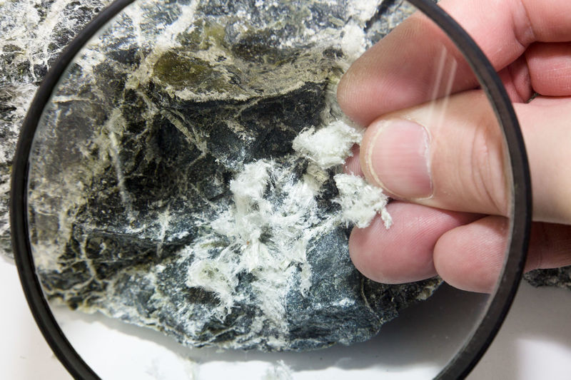 Hand with magnifying glass examines asbestos fibers in stone.