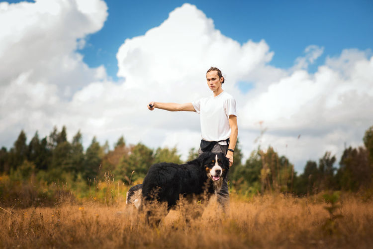 Man training dog on grassy land against cloudy sky
