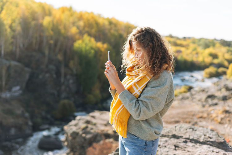 Young woman with curly hair taking photo on mobile phone on view background of mountains and river