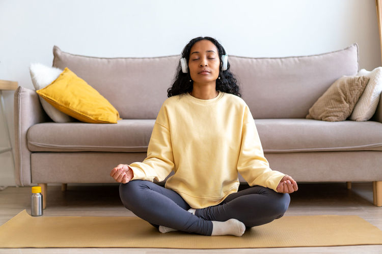 Young african-american female in headphone listening to music while meditating on mat in living room