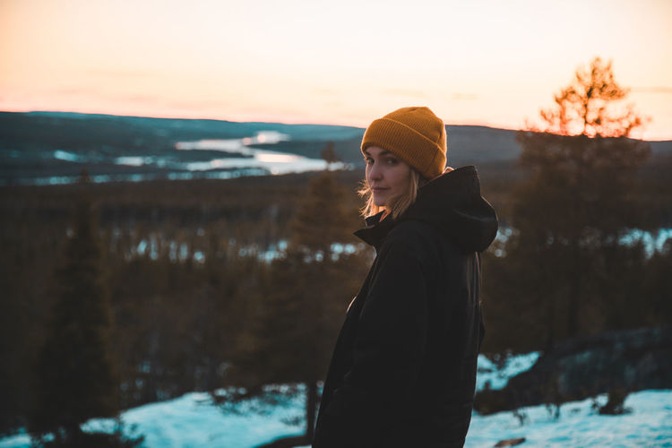Side view of calm female adventurer in warm outerwear standing on hill in evergreen woods and looking at camera on background of vivid orange sundown