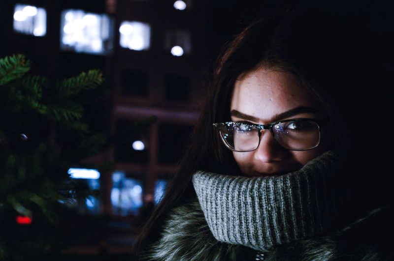 Close-up portrait of young woman in warm clothing standing against building at night