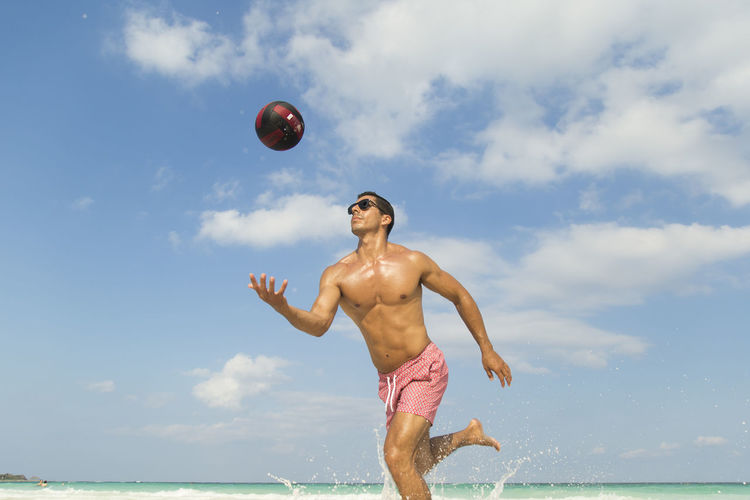 Shirtless man playing volleyball at beach against sky