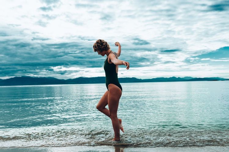 Side view of young woman in swimwear walking at lakeshore against cloudy sky