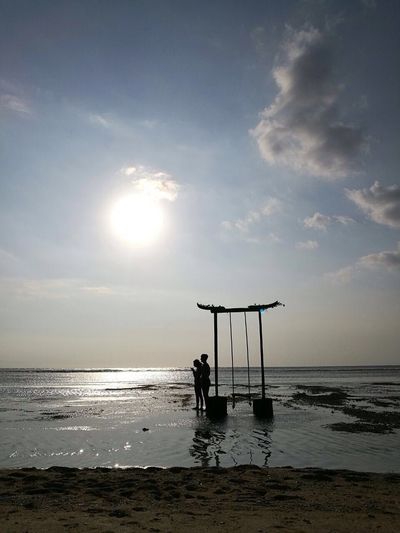Couple standing by swing on beach against sky during sunset