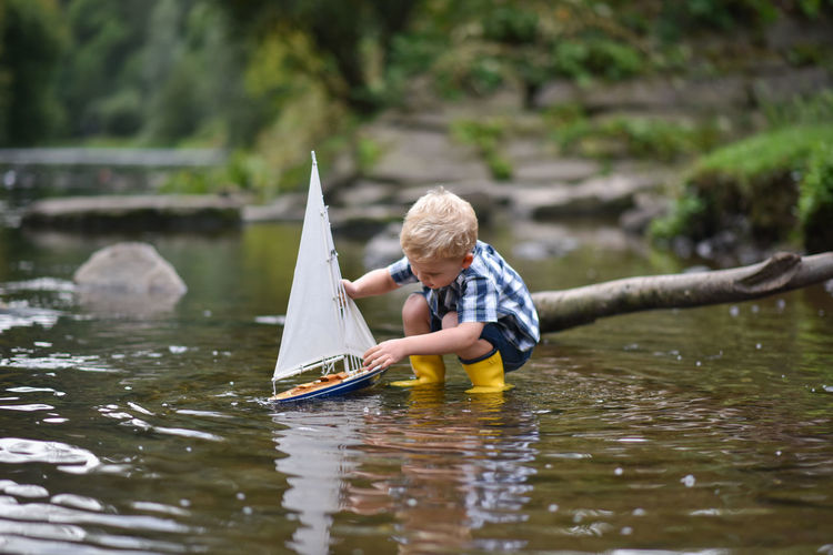 Happy little boy in yellow rain boots playing with ship boat in lake on spring or autumn day.
