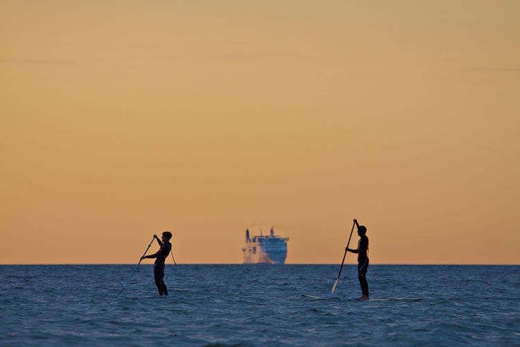 People paddleboarding on sea against sky during sunset