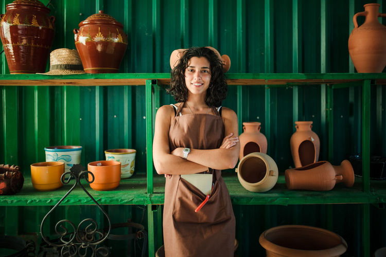 Portrait of smiling young woman standing against earthenware in workshop