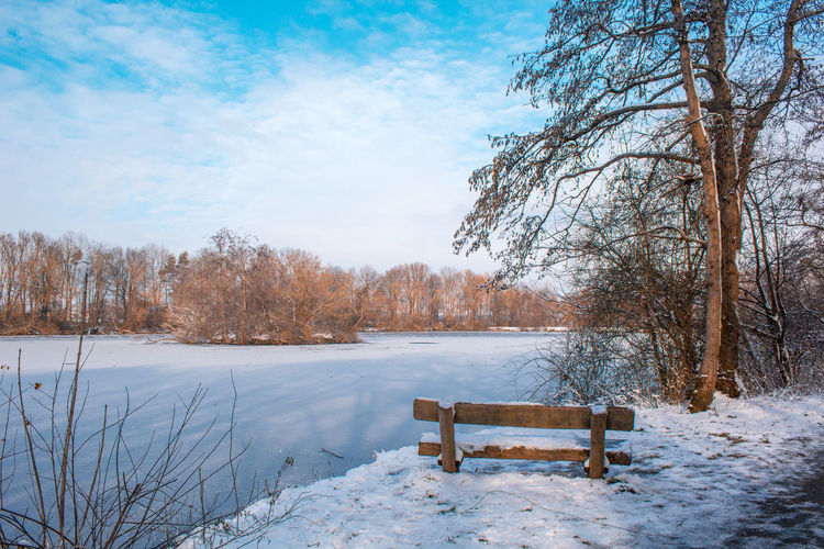 Bench by frozen lake against sky during winter