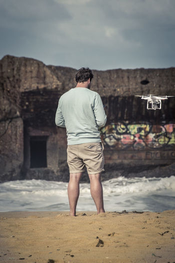 Rear view of man with drone at shore against sky