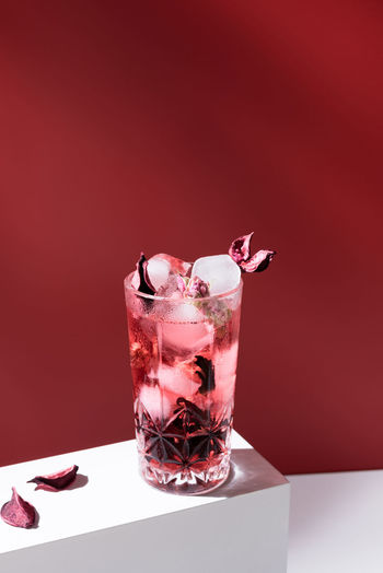 Composition of glass filled with pink gin tonic and ice placed on white table against red background