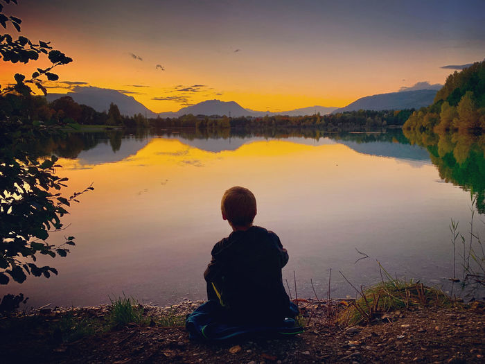 Rear view of boy sitting by lake against sky during sunset