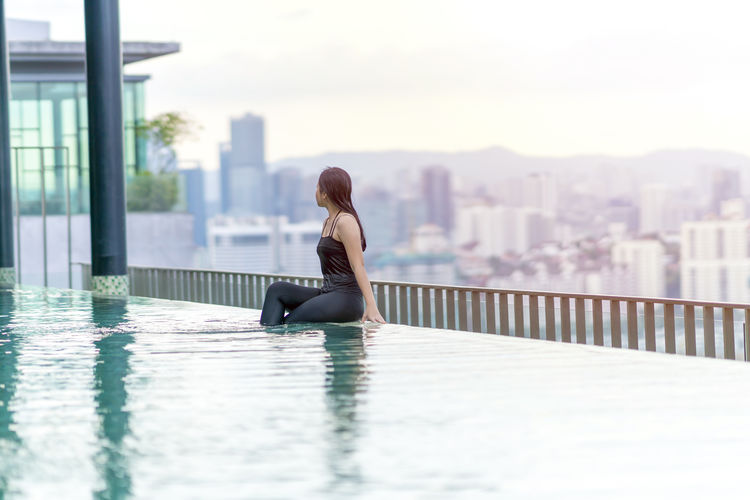 Woman sitting by swimming pool in city against sky