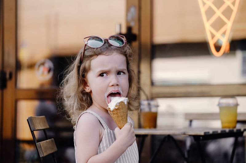 Toddler child eating cone ice cream outside near cafe. serious blonde caucasian girl licking frozen