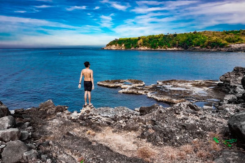 Rear view of shirtless mid adult man looking at sea while standing on rock against blue sky
