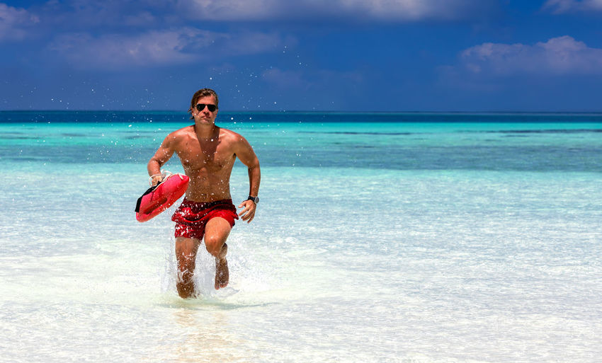 Shirtless lifeguard running at beach against sky