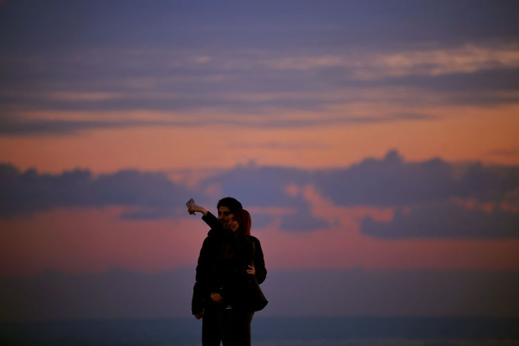 Couple taking selfie while standing against sky during sunset