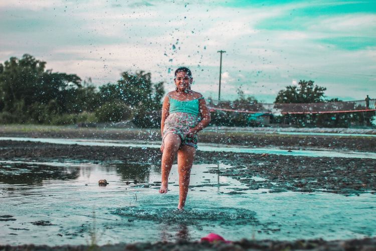 Full length of girl splashing water while standing in puddle against sky