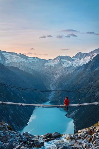 Man sitting on footbridge over lake against mountains