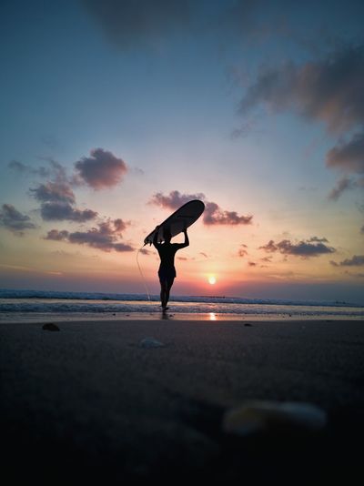 Silhouette man carrying surfboard on head at beach against sky during sunset