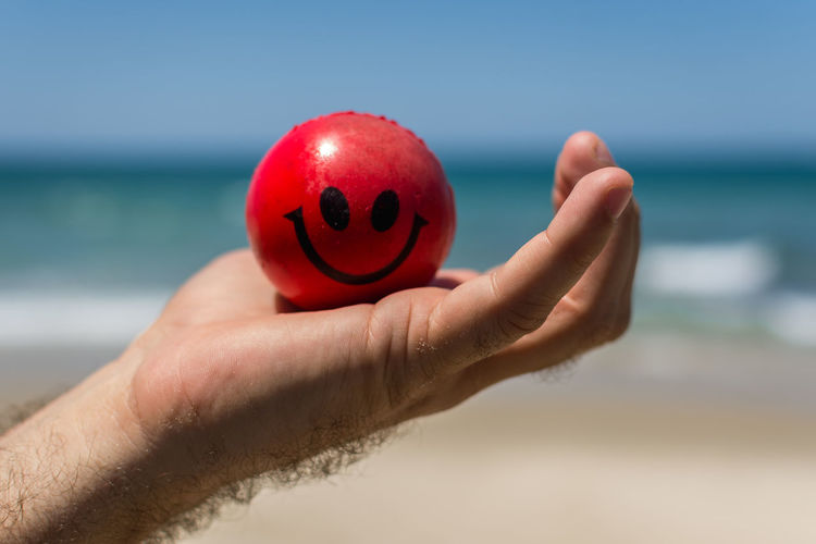 Cropped hand holding stress ball with smiley face at beach on sunny day