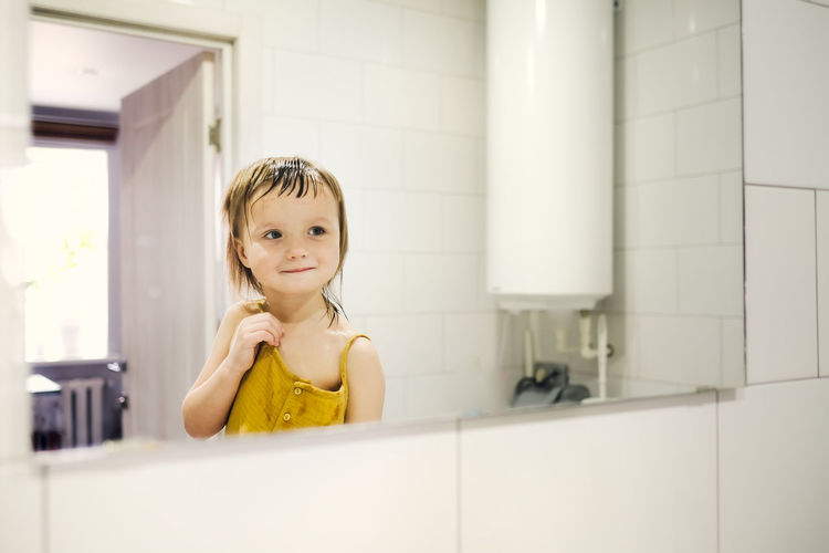 Child in yellow jumpsuit combs his own in front of mirror in bathroom