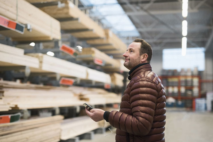 Customer looking at wooden planks on shelves while holding smart phone in hardware store warehouse