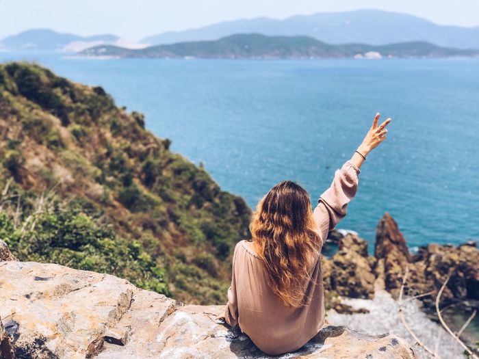 Rear view of woman showing peace sign while sitting on cliff by sea