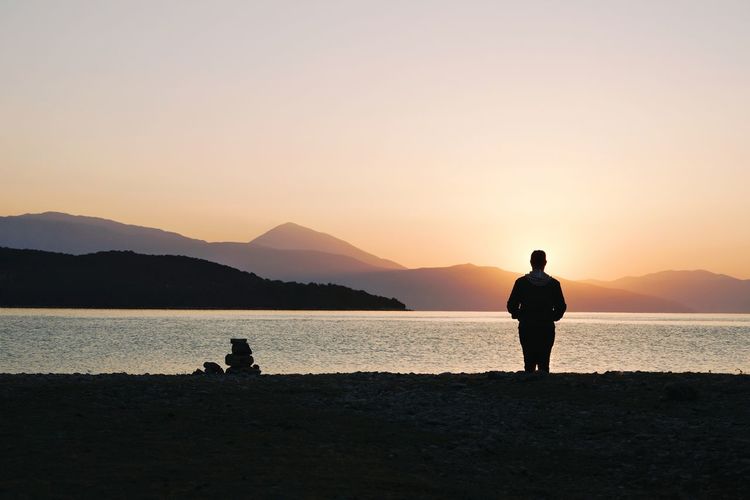 Rear view of man standing at beach against sky during sunset