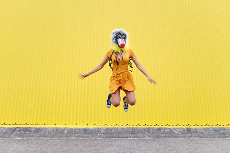 Front view of a playful woman with a mandrill mask jumping on a yellow background.