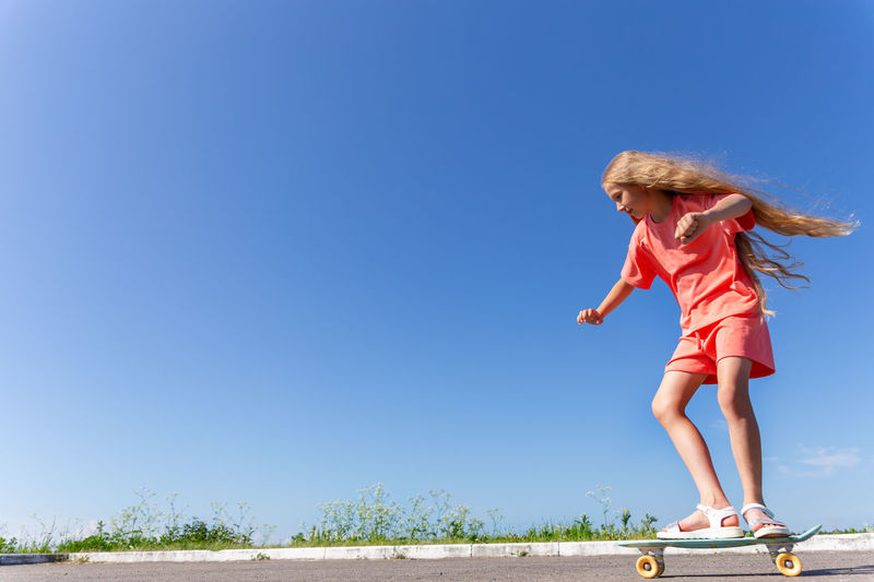 A girl is riding a skateboard with fluttering blond hair against a blue sky.
