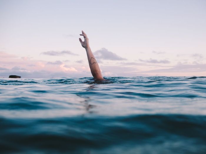 Cropped image of woman hand drowning in sea against sky