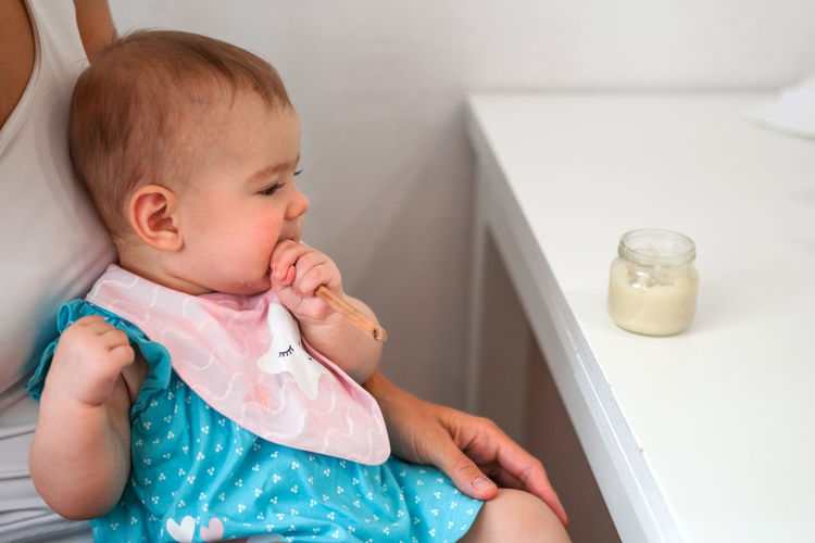 Baby girl holds wooden spoon in hand and tries to eat herself vegetable puree from glass jar.