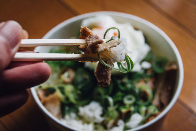 Cropped hand of person holding food in chopsticks against bowl