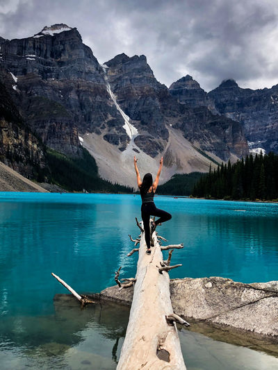 A woman does a tree pose in a log over the eater , facing the mountains. 