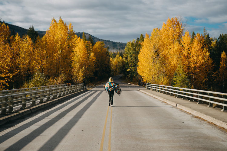 Rear view of woman walking on road against trees and sky during autumn