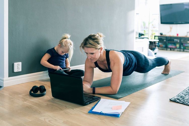 Mother looking at laptop and doing planks while daughter using smart phone in living room