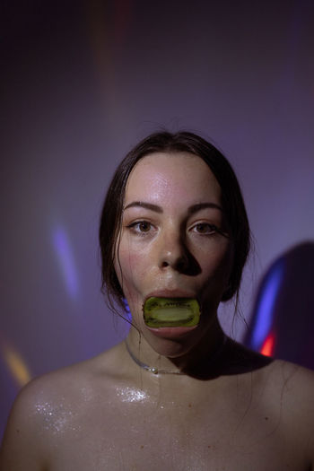 Portrait of young woman with kiwi fruit in mouth against wall