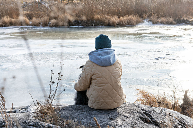 Young woman sitting on stone hugging dog looking at river covered with first ice in fall or winter