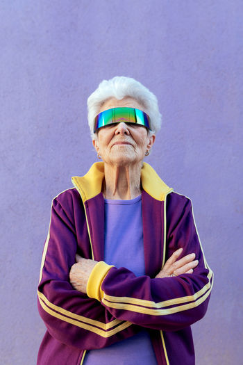 Elderly female athlete with folded arms and gray hair in sportswear and blindfold on violet background
