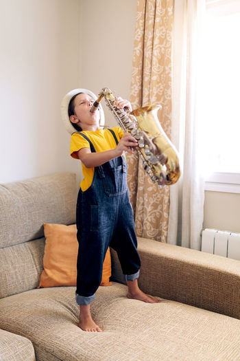 Barefoot child with closed eyes playing saxophone while standing on couch at home in daytime