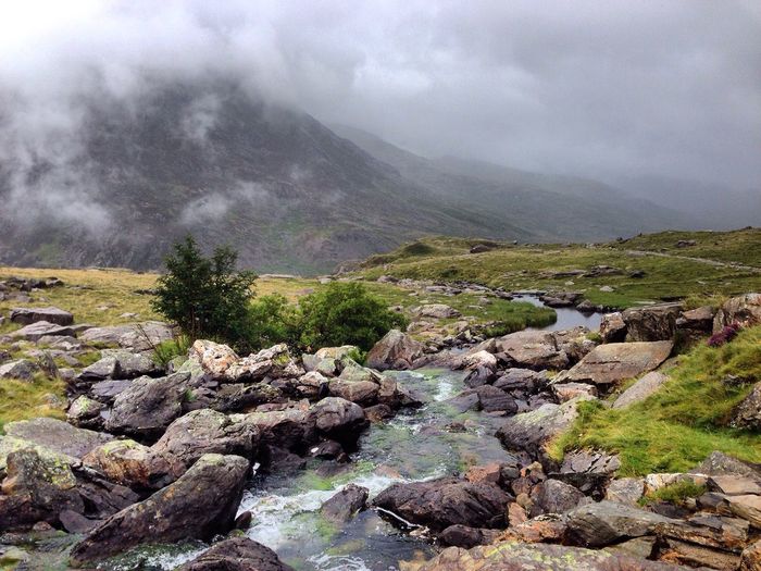 Stream flowing through rocks on hill against mountain covered with clouds