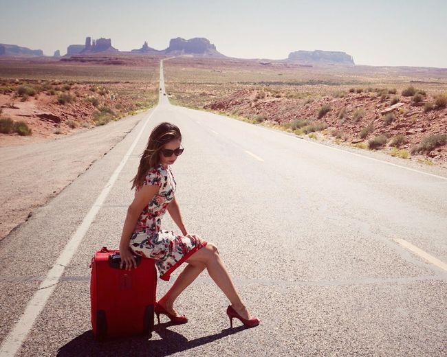 Full length of beautiful woman with luggage on road leading towards monument valley during sunny day