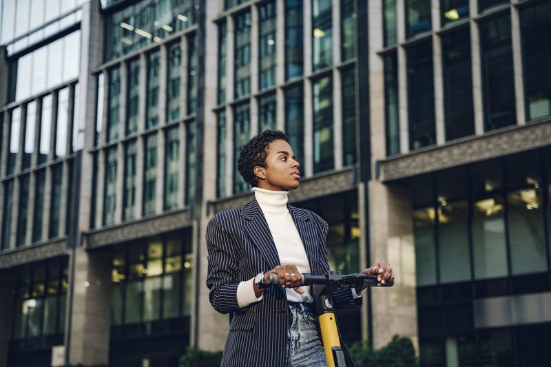 Thoughtful young businesswoman with electric push scooter against modern building in city