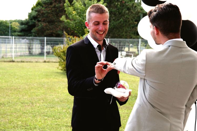 Cheerful man putting ring on boyfriend at park during wedding ceremony