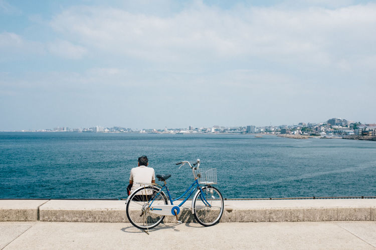 Rear view of man with bicycle on promenade by sea against cloudy sky