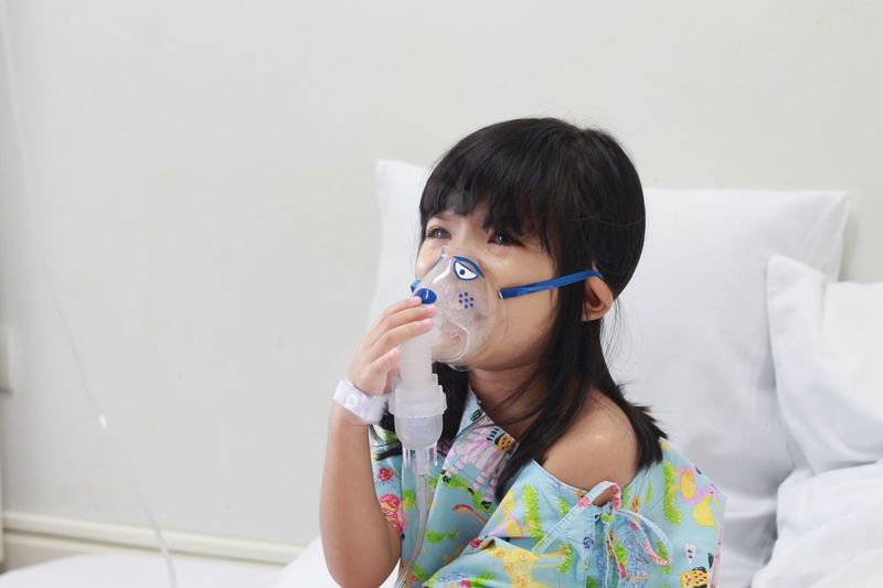Girl breathing with oxygen mask while sitting on bed in hospital
