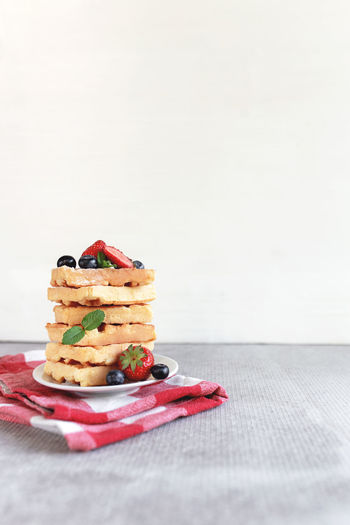 Stack of waffles on a white plate on the towel and table with blueberry, chopped strawberry and mint
