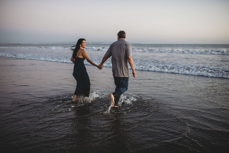 Laughing barefoot couple wading into the ocean in their street clothes
